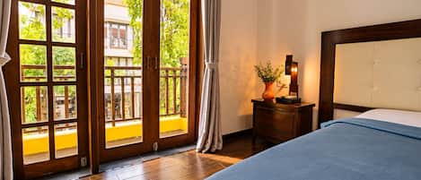 Habitación de hotel con piso de madera, cama con cubrecama azul, mesita de noche con un florero y una lámpara, y vista a un balcón exterior a través de una puerta de vidrio.