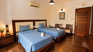 A hotel room with two beds, a wooden floor, a wall-mounted air conditioner, a bedside table with a lamp, a chair, and a door.