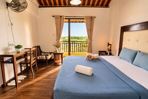 A hotel room with a bed, a desk, a chair, a fan, and a view of greenery through the window. - Flower’s House Hoi An (Da Nang)