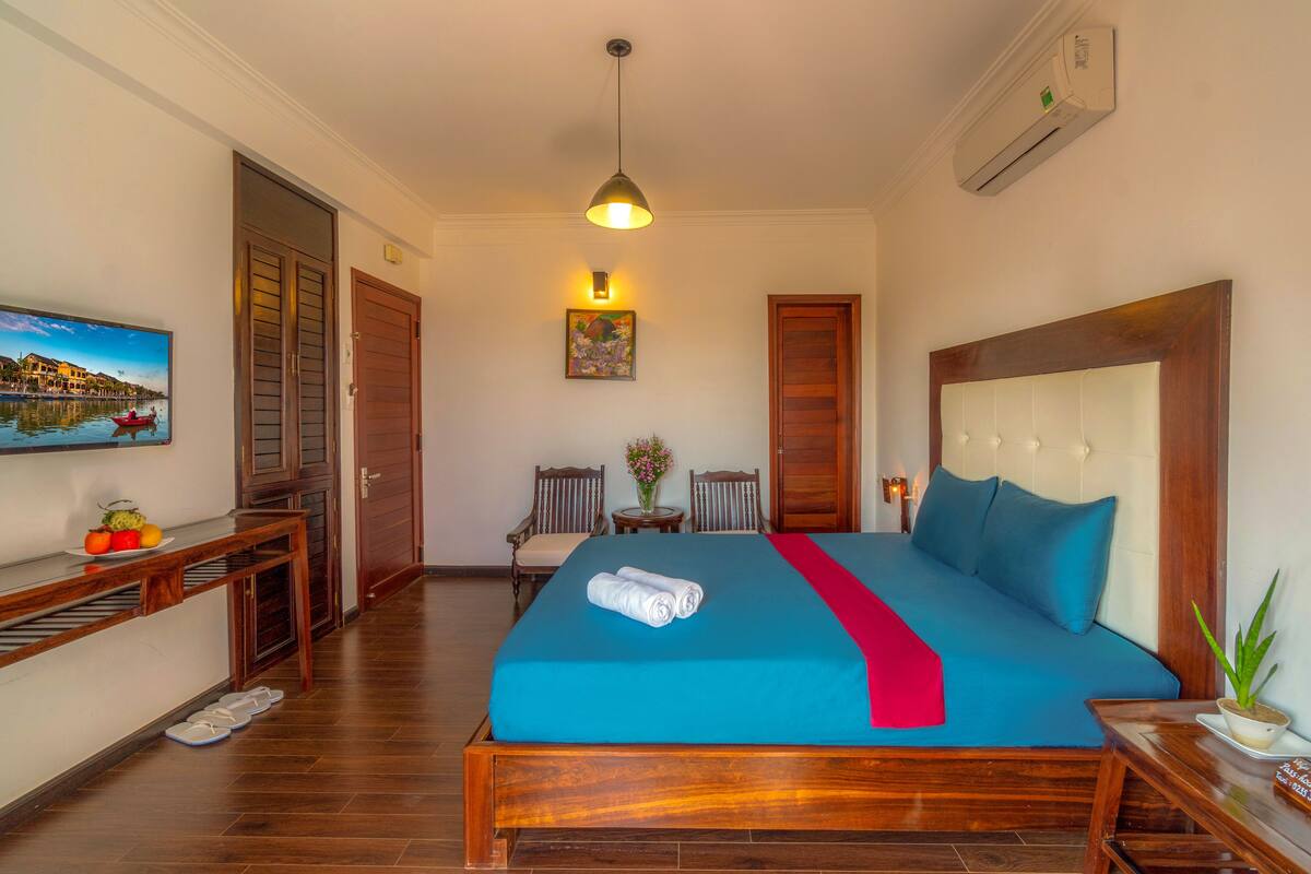 A hotel room with a wooden bed, a television on the wall, a small table with a vase, and a wooden bench.