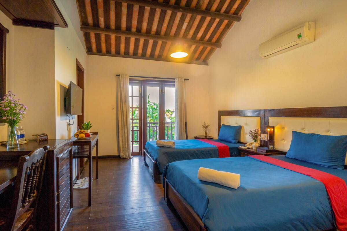 A hotel room with two beds, a desk, a chair, a TV, and a balcony with a view of greenery.