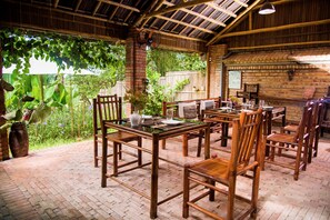 An outdoor dining area with bamboo furniture and brick flooring, surrounded by lush greenery. - Calm House Hotel Hoi An (Da Nang)