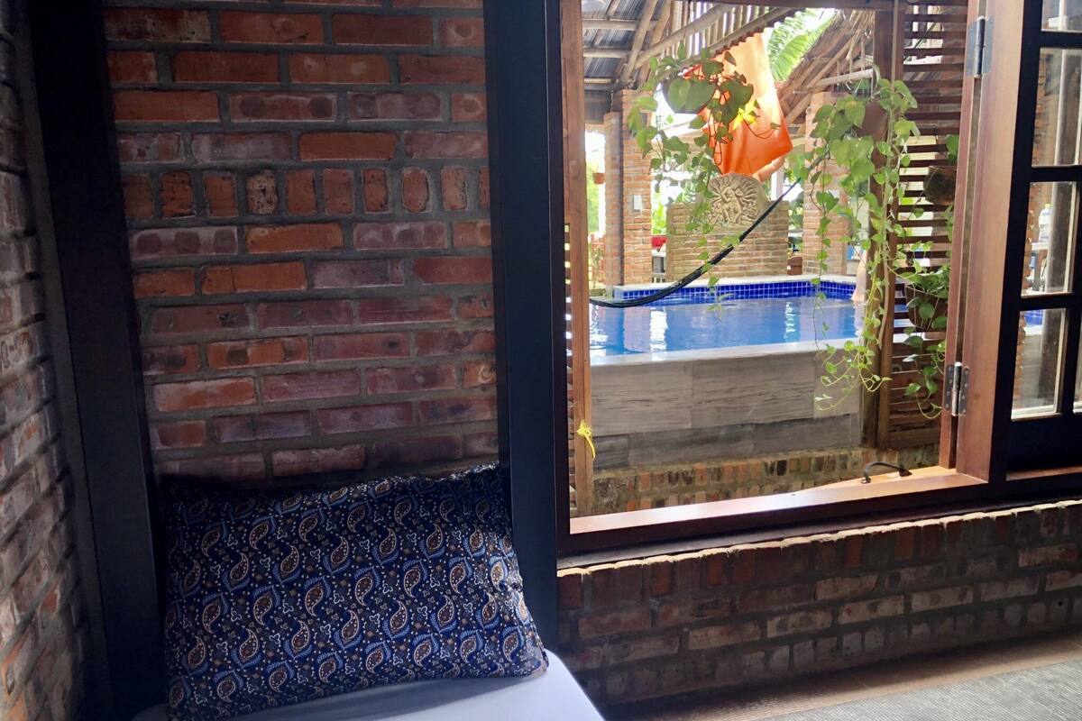 A room with a brick wall, a window with a view of a pool and plants, and a patterned cushion on a bench.