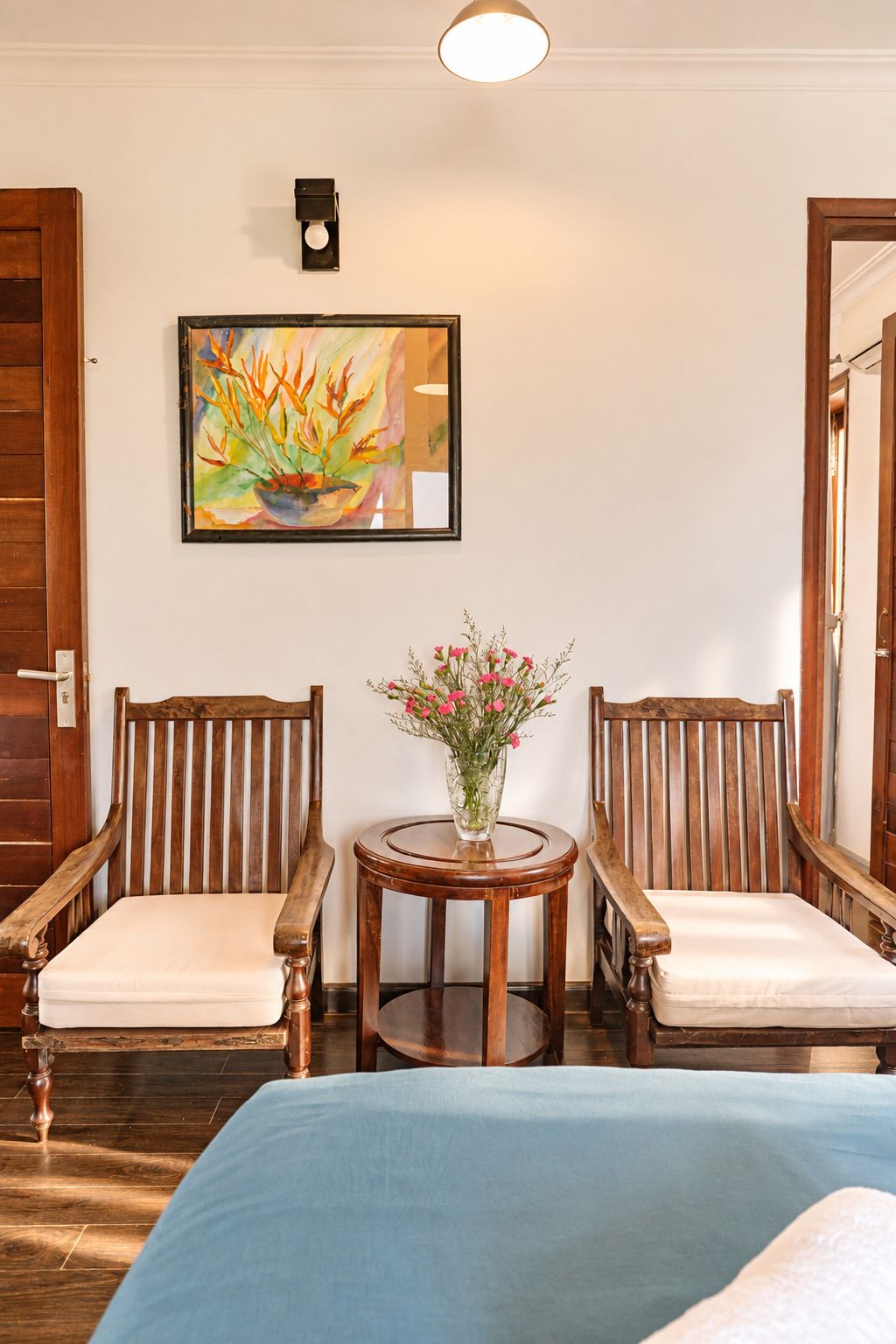 A room with two wooden chairs, a small round table with a vase of flowers, and a bed with a blue cover.
