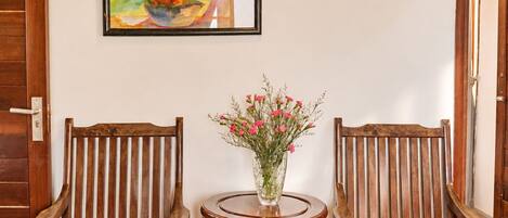 A room with two wooden chairs, a small round table with a vase of flowers, and a bed with a blue cover.