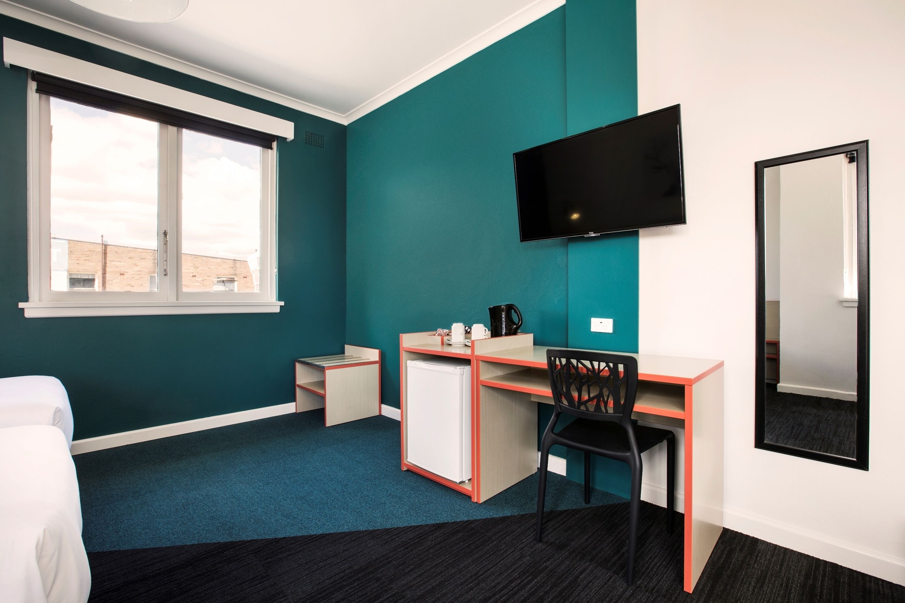 A hotel room with teal walls, a desk with a chair, a bed, a TV, and a small refrigerator.