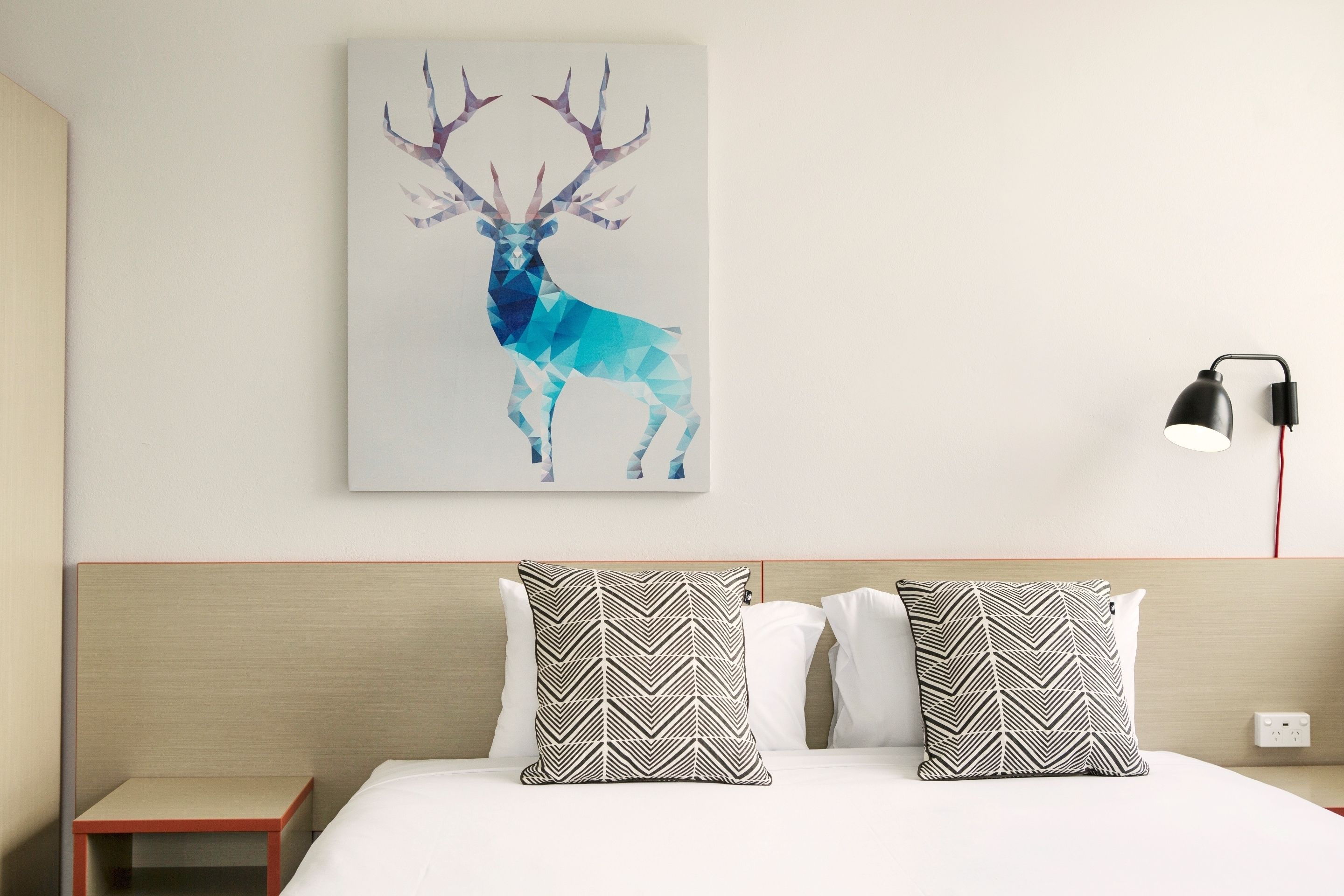 A hotel room with a bed, two patterned pillows, a wall-mounted artwork of a deer, and a bedside lamp.