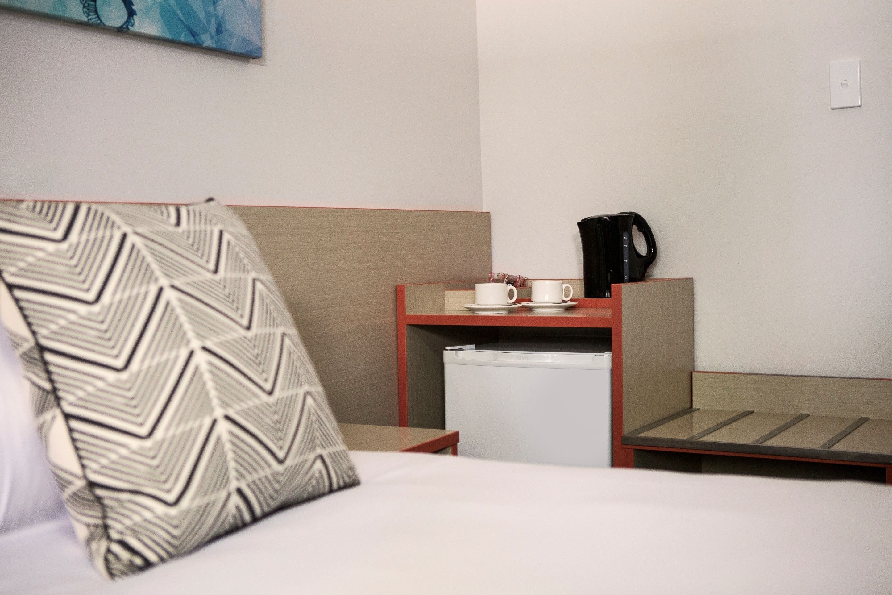 A hotel room with a bed, a bedside table with a coffee maker, a small refrigerator, and a wall-mounted painting.