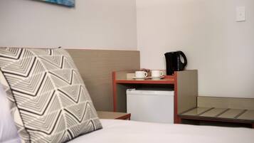 A hotel room with a bed, a bedside table with a coffee maker, a small refrigerator, and a wall-mounted painting.