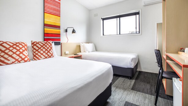 A hotel room with two beds, a desk, a chair, a window, and a wall art piece. - Villawood Hotel (Villawood)