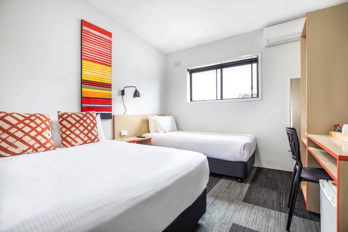 Habitación de hotel con dos camas, un escritorio, una silla, una ventana y una obra de arte en la pared.
