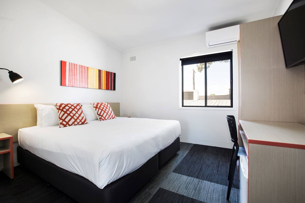Habitación de hotel con una cama grande, un escritorio, una silla, un televisor, una ventana y un cuadro en la pared.