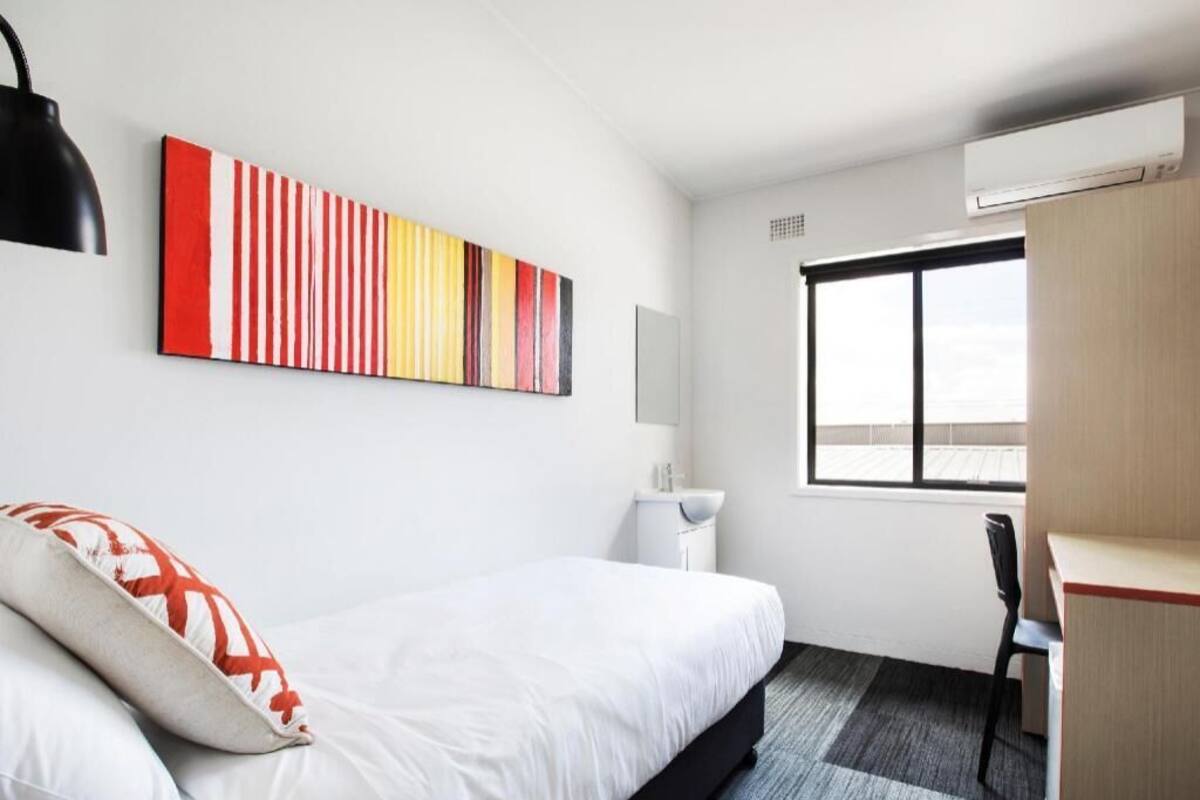 Una habitación de hotel con cama, escritorio, silla, ventana y aire acondicionado de pared.