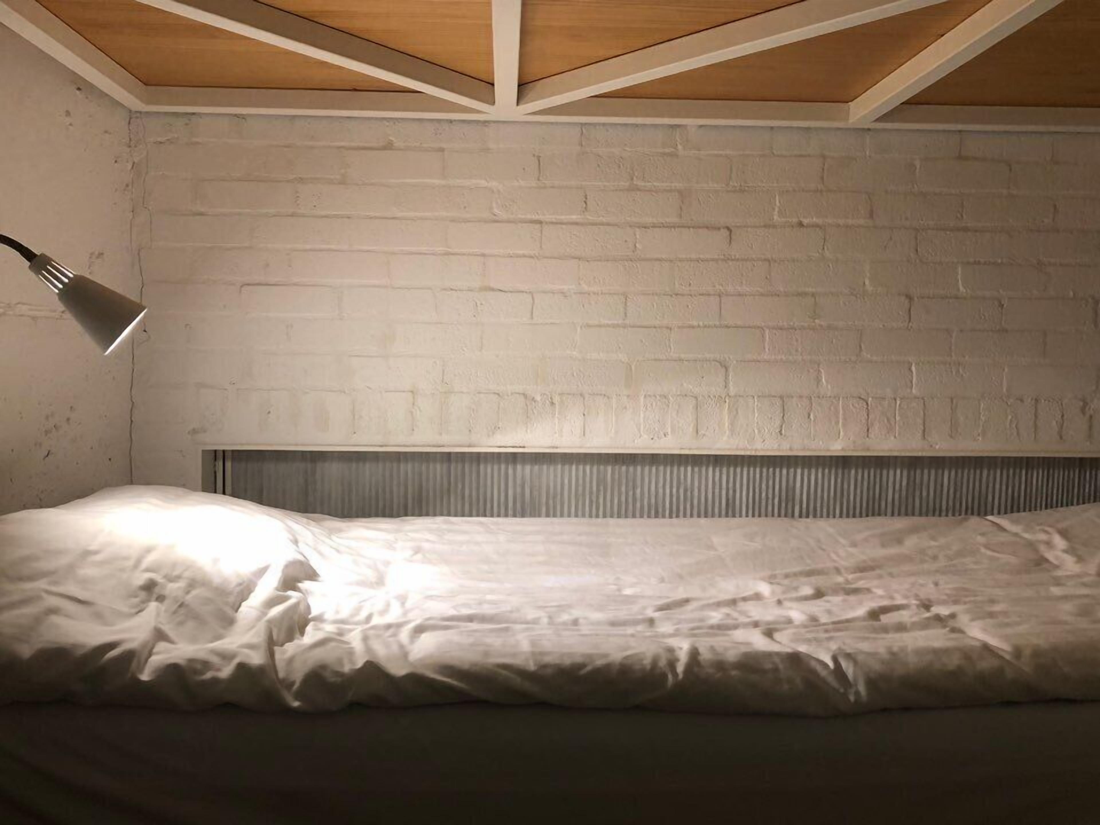 A bedroom with a white brick wall, a slanted ceiling, and a single bedside lamp.