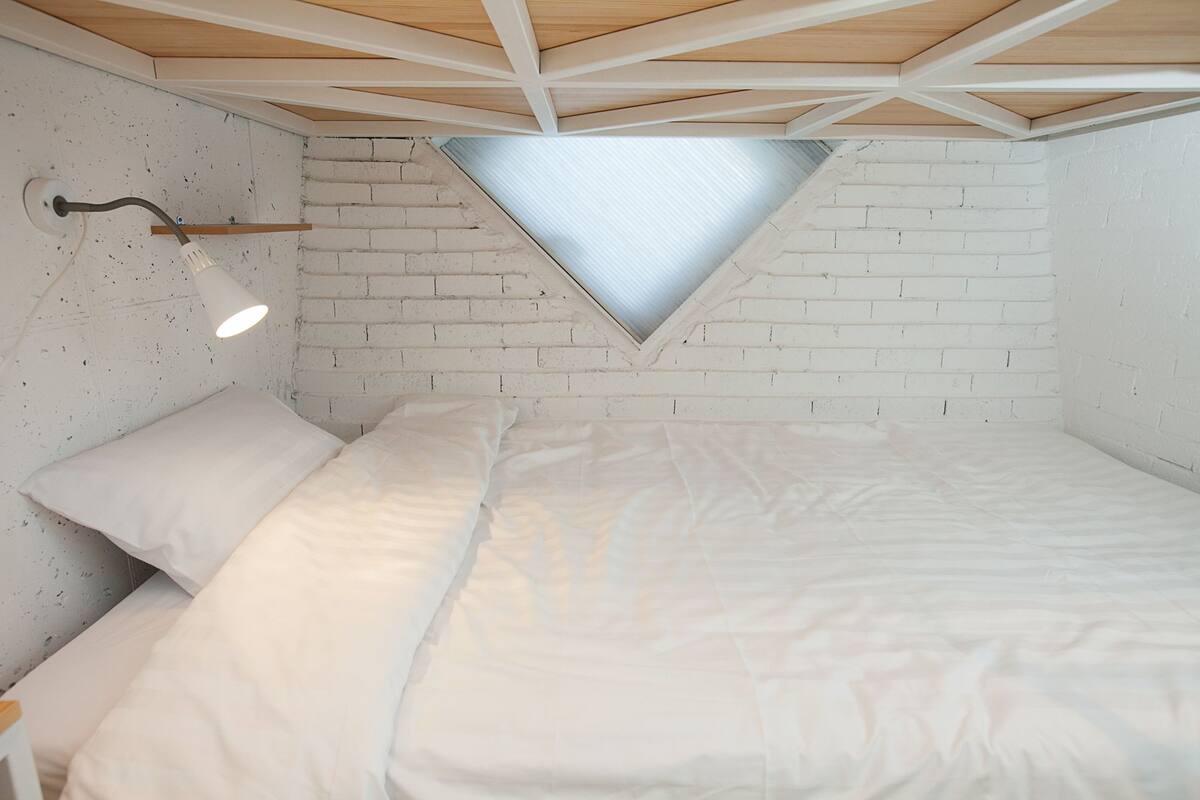 Une chambre avec un plafond en pente, un puits de lumière, un lit avec une literie blanche et une table de chevet avec une lampe.