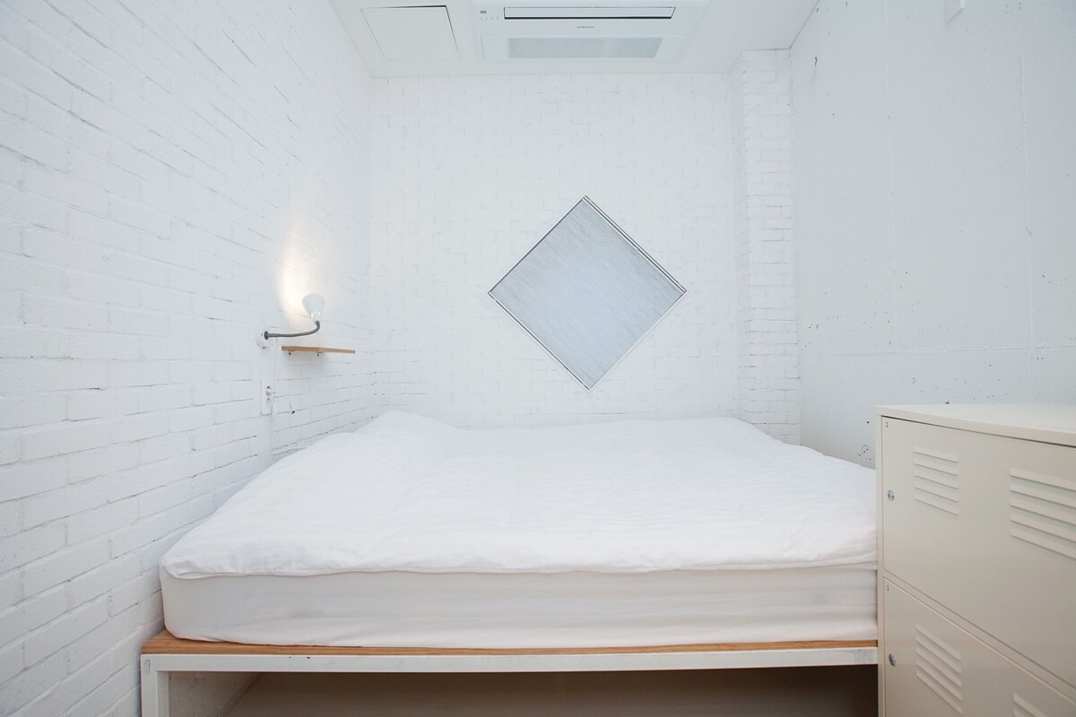 A small, white-walled room with a single bed, a wall-mounted light, and a cabinet.