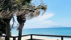 Eine Holzterrasse mit weißen Plastikstühlen und einem Tisch mit Blick auf einen Strand, Palmen und einen klaren, blauen Himmel.