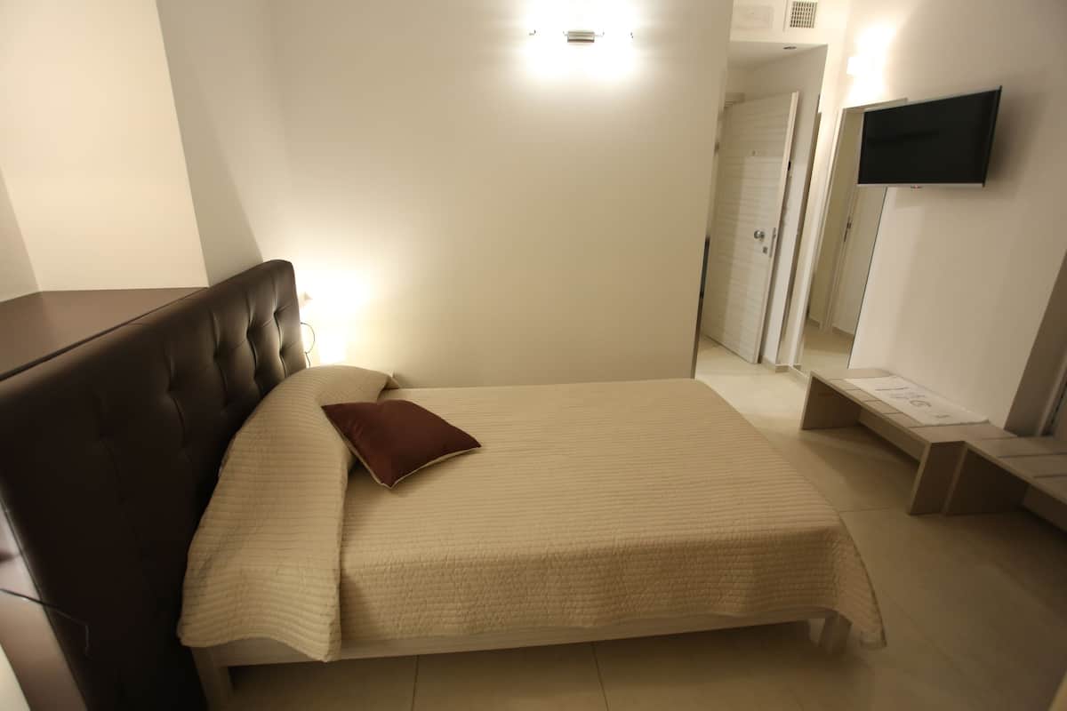Una cama bien hecha con un almohadón marrón, una mesita de noche, un televisor de pantalla plana montado en la pared y una puerta que da a otra habitación.