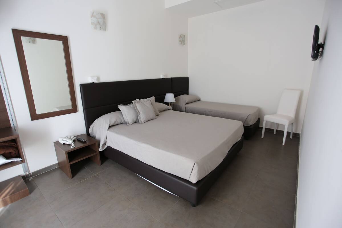 Habitación de hotel con una cama grande, otra más pequeña, una mesita de noche con teléfono, una silla y un televisor montado en la pared.