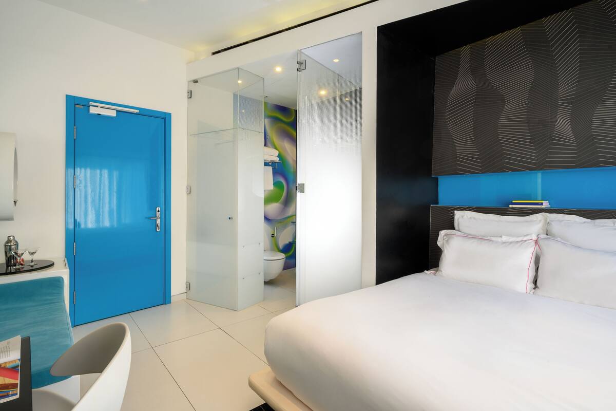Une chambre d’hôtel moderne avec un grand lit, une porte bleue, une cabine de douche en verre et une tête de lit noire.