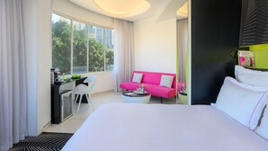 Een moderne hotelkamer met een groot bed, een roze bank, een eettafeltje en uitzicht op groen.