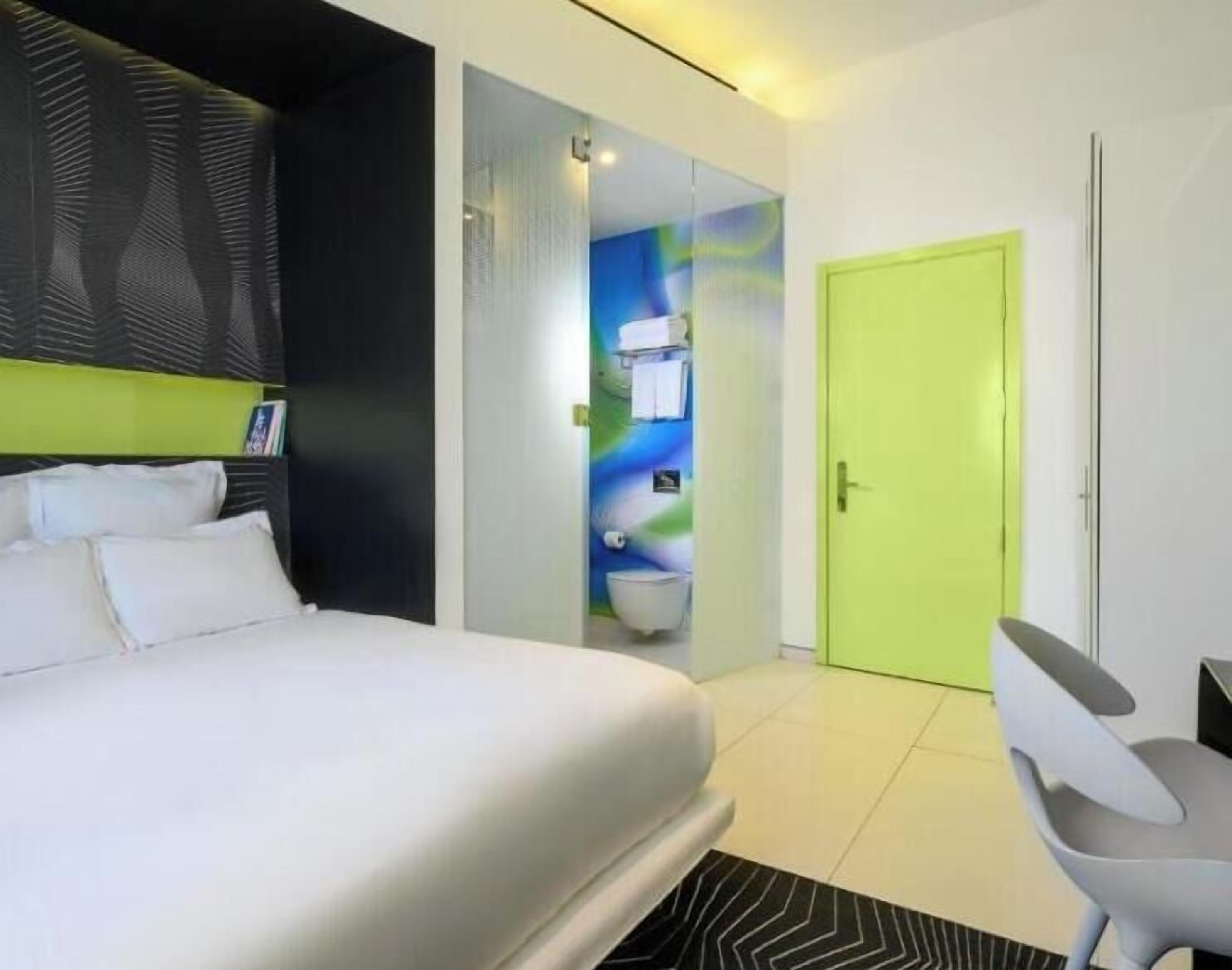 A modern hotel room with a large bed, a bright green door, and a bathroom visible through a glass partition.