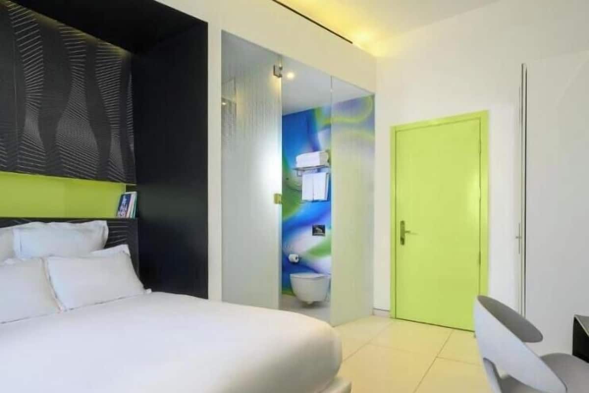 Une chambre d’hôtel moderne avec un grand lit, une porte vert vif et une salle de bain visible à travers une paroi en verre.
