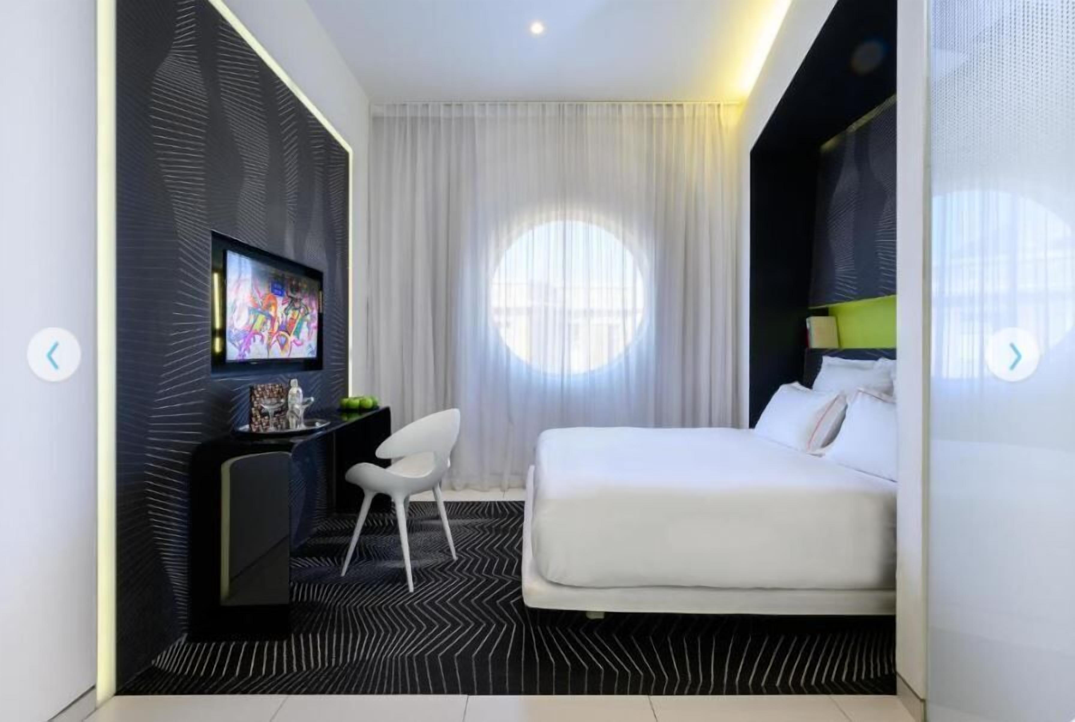 A modern hotel room with a bed, a desk with a TV, a chair, and a large window with curtains.