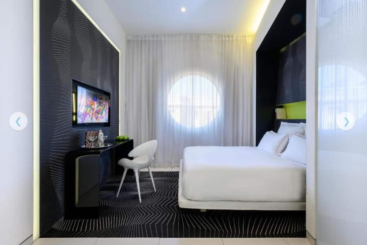 Une chambre d’hôtel moderne équipée d’un lit, d’un bureau avec une télévision, d’une chaise et d’une grande fenêtre avec des rideaux.