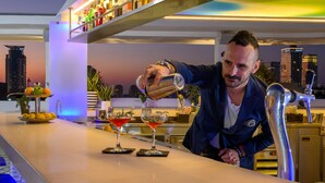 Een bar met een bartender die drankjes inschenkt, een fruitschaal en een stadsgezicht op de achtergrond.
