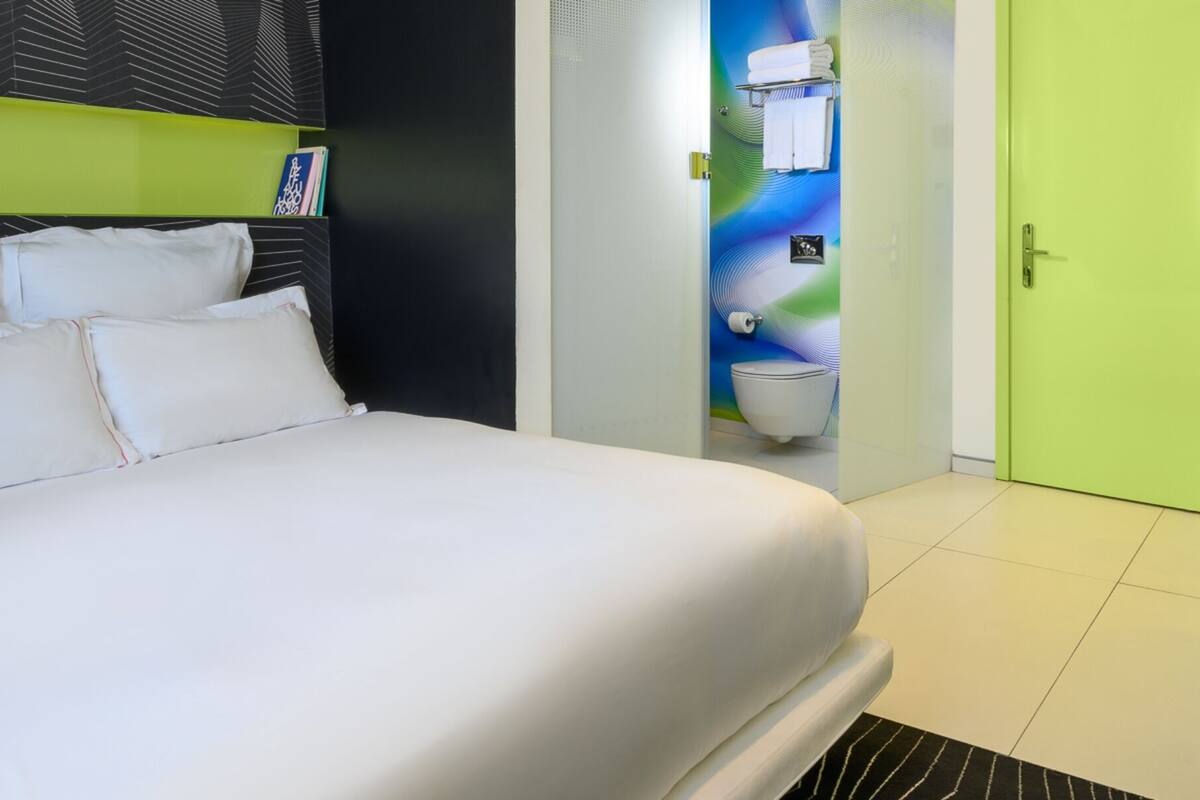 Une chambre d’hôtel moderne avec un grand lit, une salle de bain avec un mur vert et un coin salon agrémenté d’un panier de pommes.