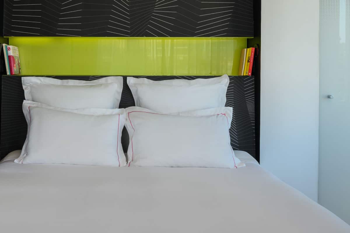 Un lit bien fait, avec du linge de lit blanc, une tête de lit noire et verte, et un petit bureau avec une chaise et des livres.