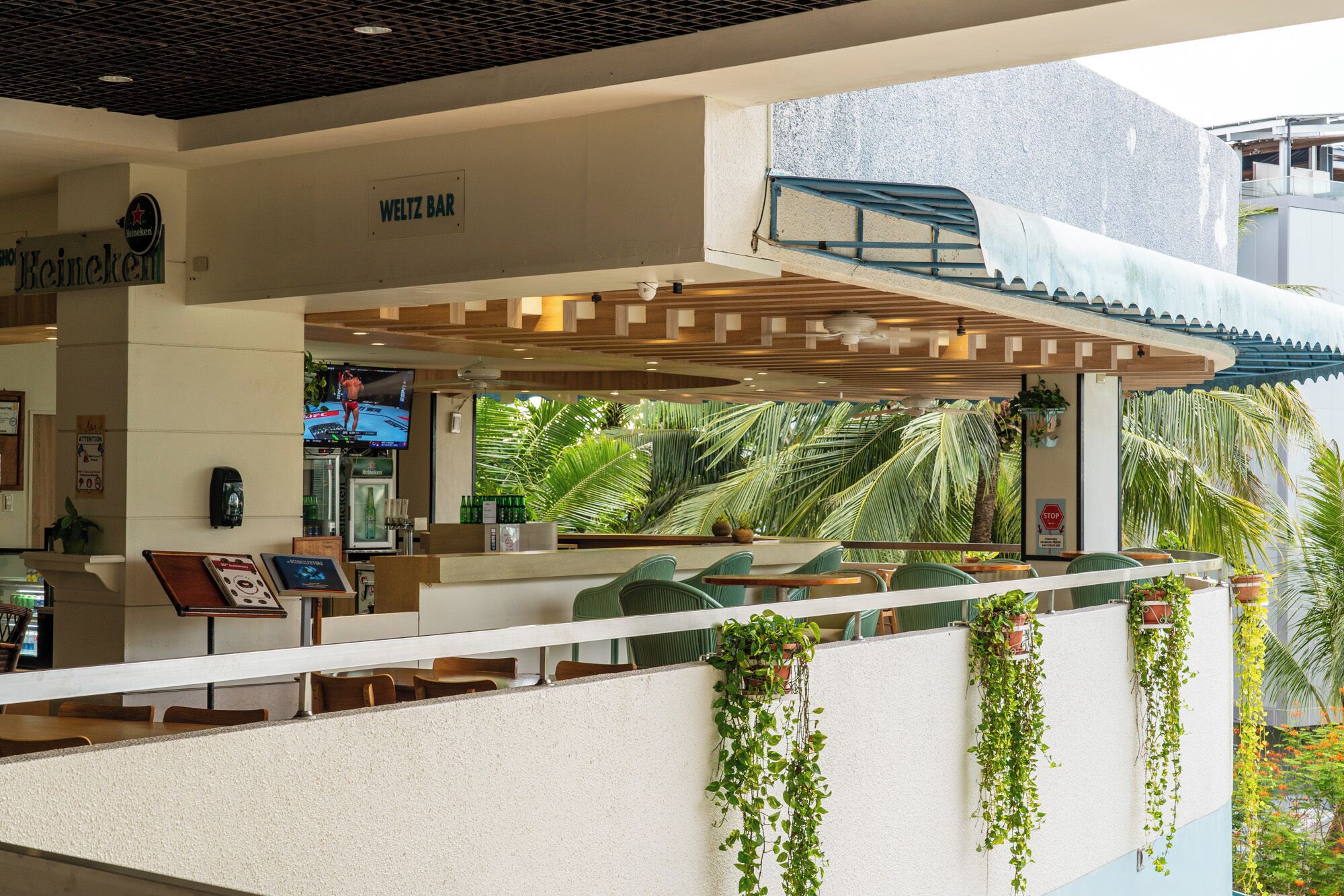 A modern bar area with a sign for 'WELTZ BAR' and a Heineken logo. There are outdoor seating with plants and a view of palm trees.