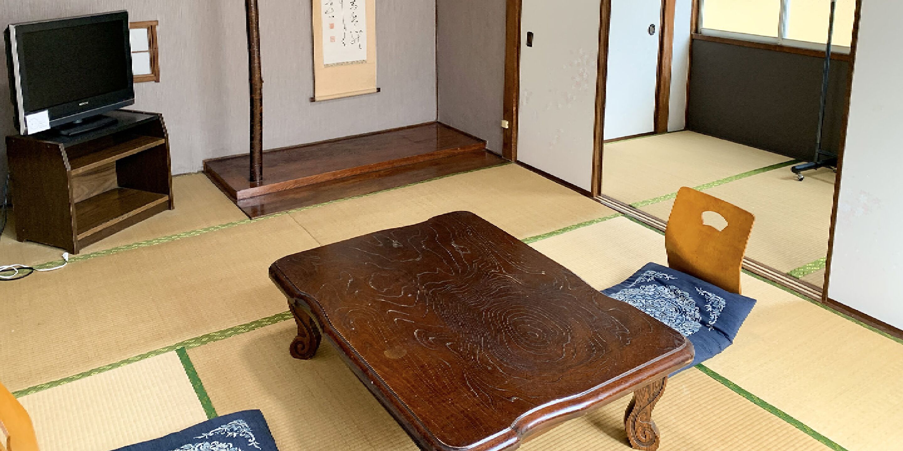 A traditional Japanese room with tatami flooring, a low wooden table, a television on a stand, and a sliding door.