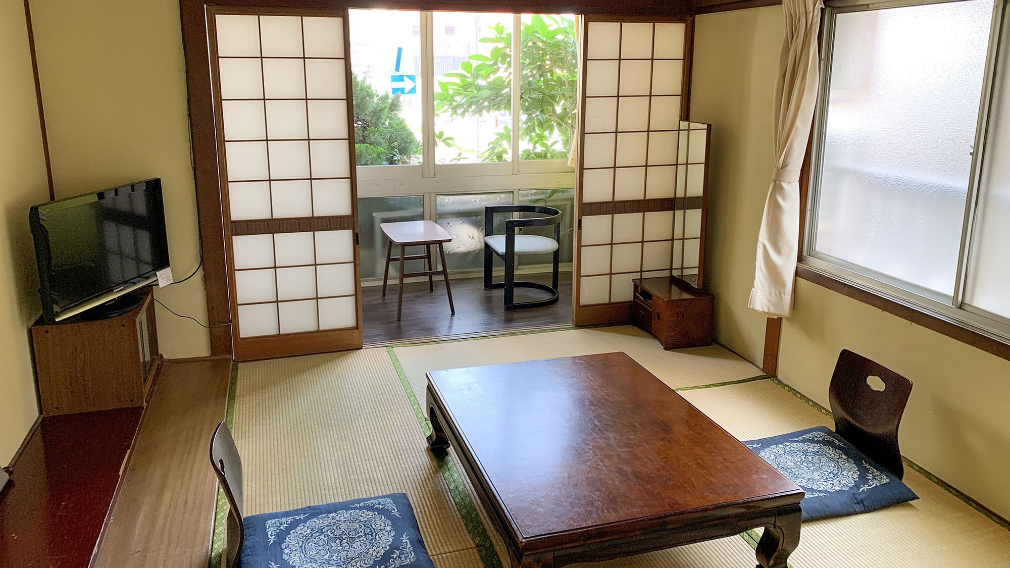 A traditional Japanese room with tatami flooring, a low table, a television on a stand, and sliding doors leading to an outdoor area.