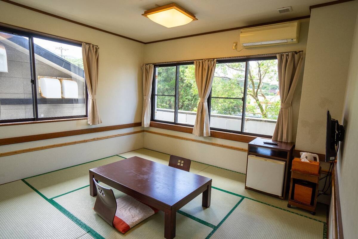 Ein Zimmer mit Tatami-Bodenbelag, einem Holztisch, einem Stuhl, einer kleinen Kommode und einem Fenster mit Vorhängen.