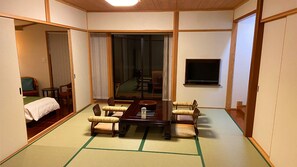 A traditional Japanese-style room with tatami flooring, a low table with cushions, and a television on the wall. - Yakushima Jomon No Yado Manten (Yakushima)