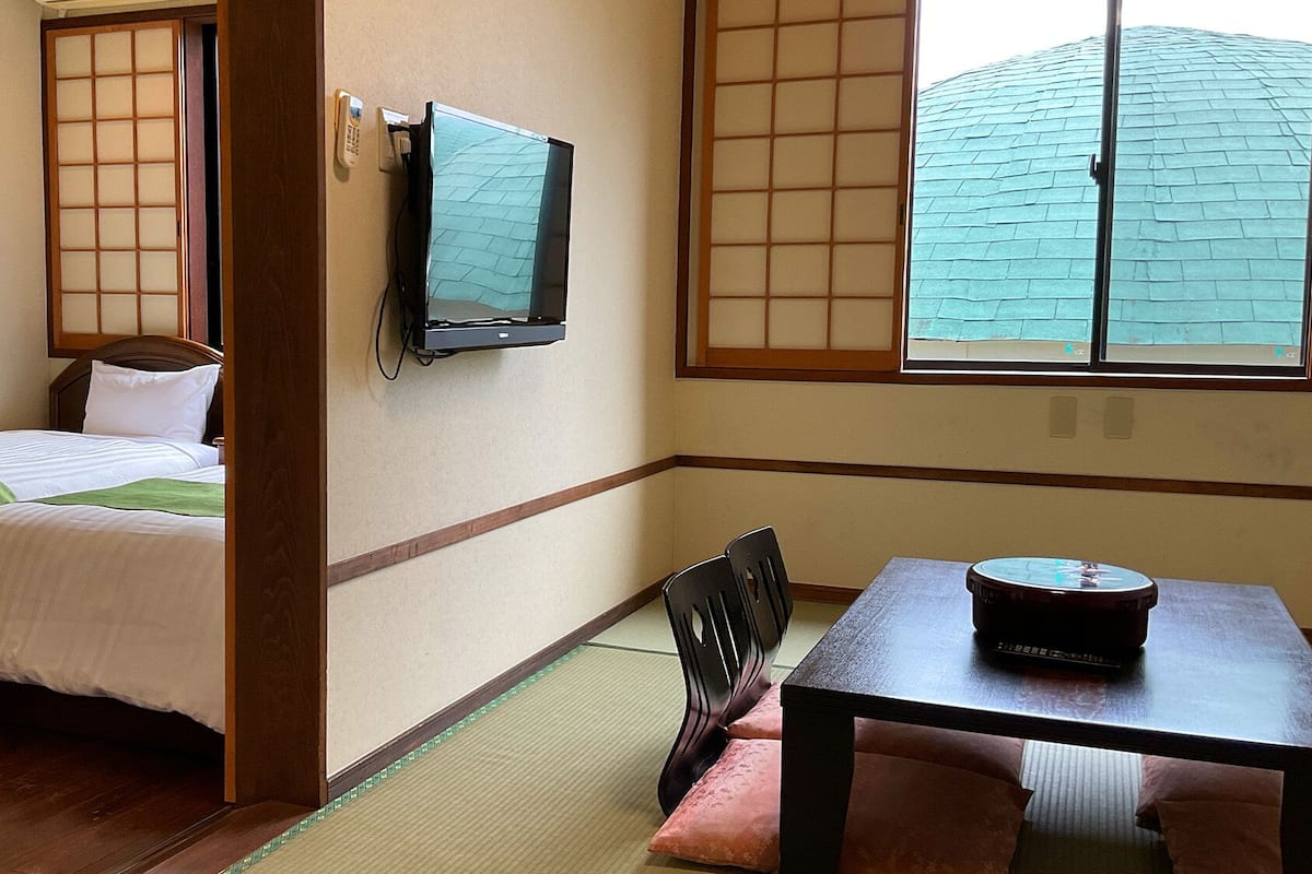 Ein traditioneller japanischer Raum mit Tatami-Bodenbelag, einem Bett mit grüner Decke, einem an der Wand montierten Fernseher und einem Tisch mit Fernbedienung.