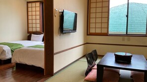 A traditional Japanese-style room with tatami flooring, a bed with a green blanket, a television mounted on the wall, and a table with a remote control. - Yakushima Jomon No Yado Manten (Yakushima)
