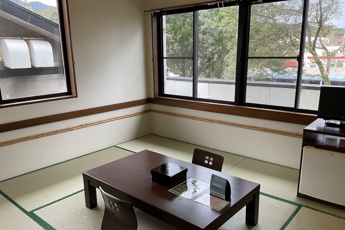 Ein Zimmer mit Tatami-Bodenbelag, einem Holztisch, einem Stuhl und einem kleinen Kühlschrank.