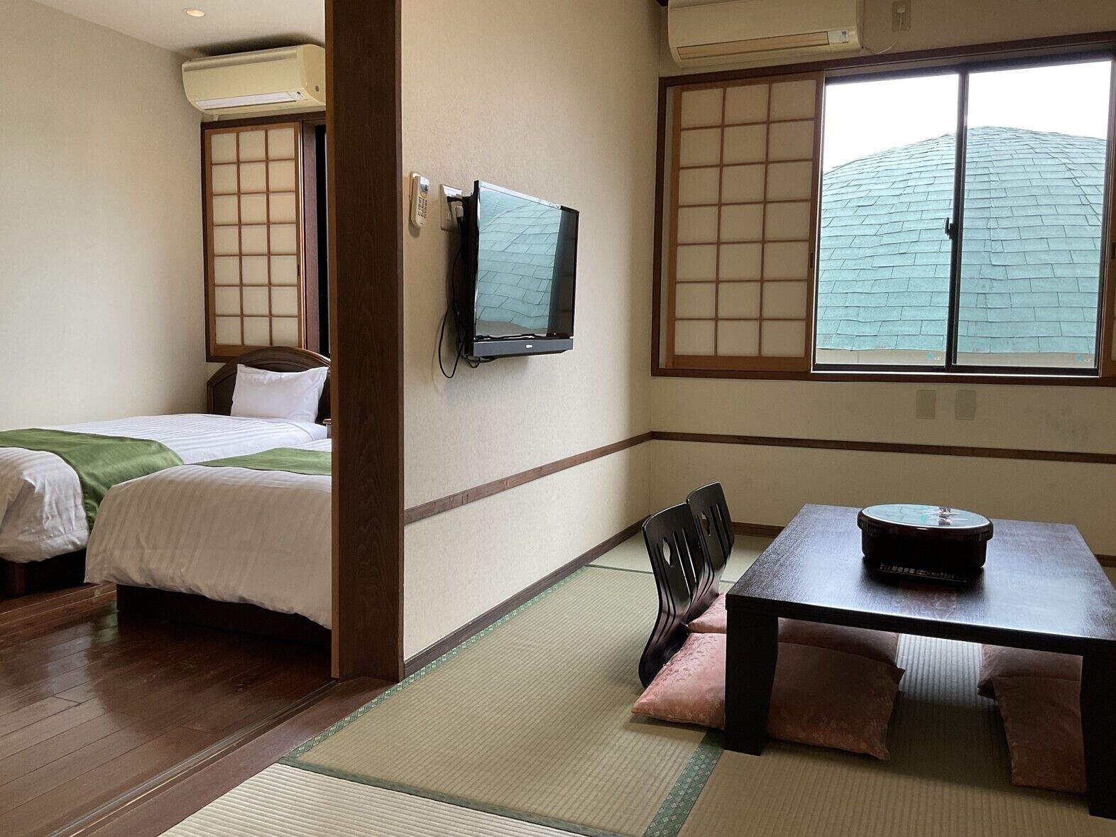 Une chambre d’hôtel avec un lit, une table avec une radio, une télévision et une fenêtre donnant sur un immeuble.