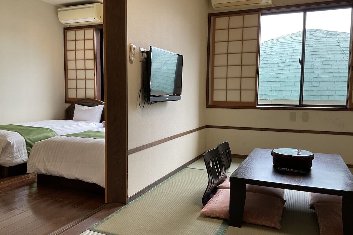 Ein Hotelzimmer mit einem Bett, einem Tisch mit Radio, einem Fernseher und einem Fenster mit Blick auf ein Gebäude.