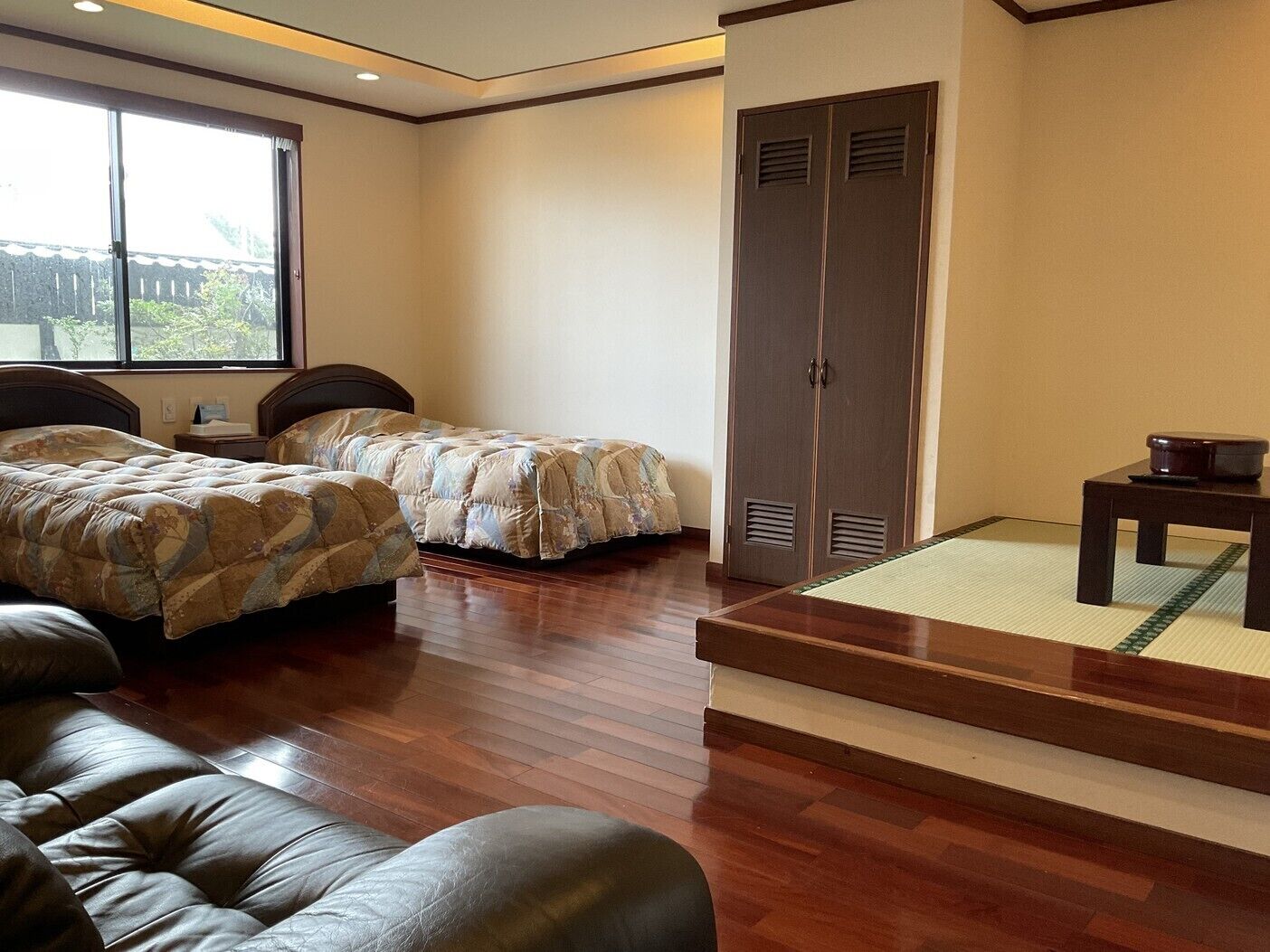 A hotel room with two beds, a sofa, a small table, a wardrobe, and a view through a window.