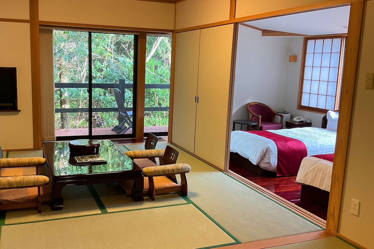 Ein traditioneller japanischer Raum mit Tatami-Böden, einem niedrigen Tisch mit Kissen, einem Fernseher und einer Schiebetür zu einem Schlafzimmer mit Bett und Schreibtisch.
