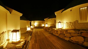 Un sendero de madera que conduce a un muro de piedra, flanqueado por edificios blancos con faroles iluminados.