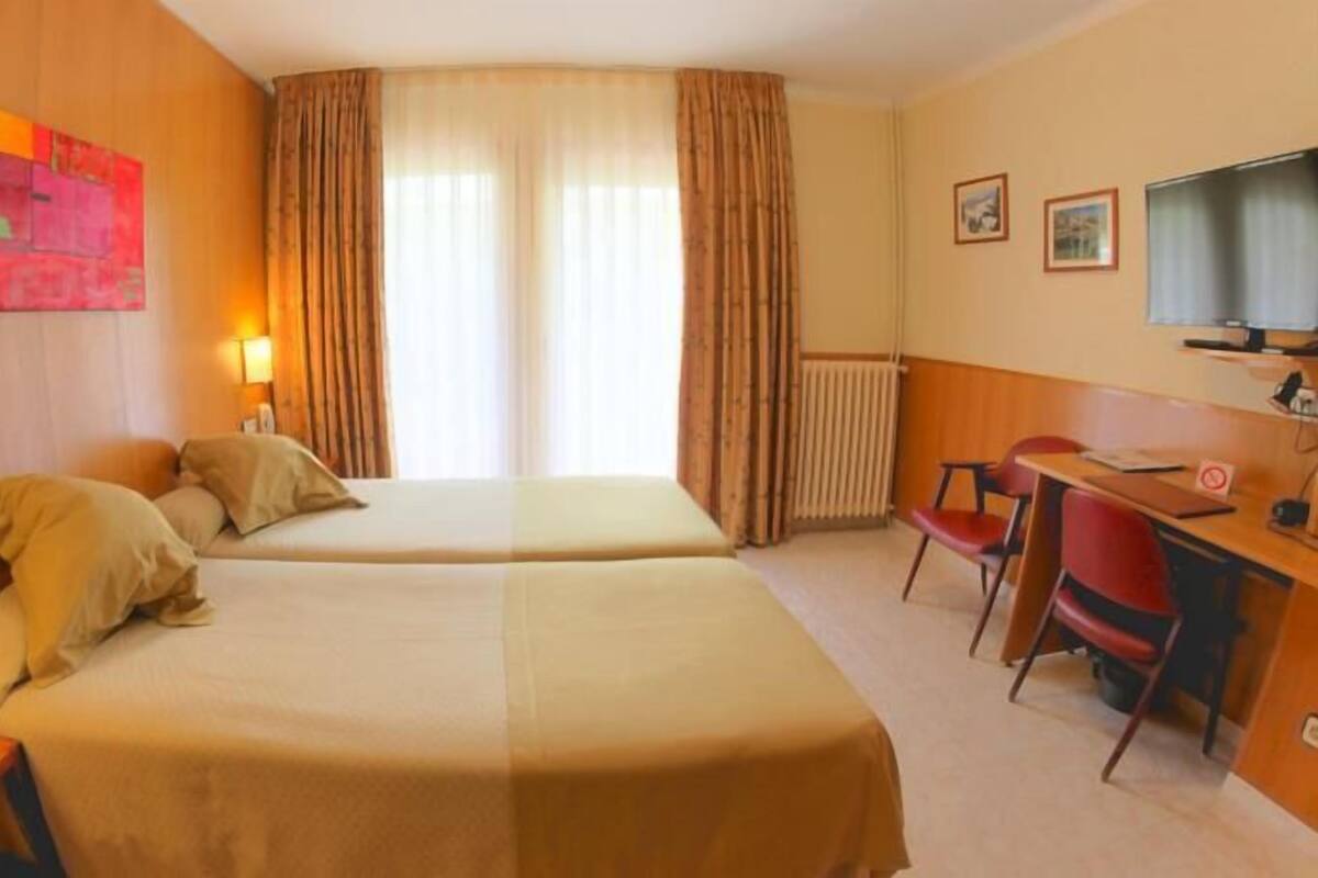 Habitación de hotel con dos camas, un escritorio, una silla, un televisor y una ventana con cortinas.