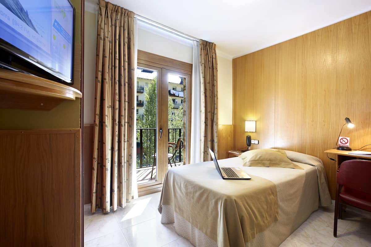 Habitación de hotel con una cama, un escritorio con una computadora portátil, un televisor y un balcón con cortinas.