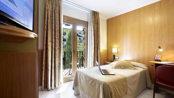 Habitación de hotel con una cama, un escritorio con una computadora portátil, un televisor y un balcón con cortinas.
