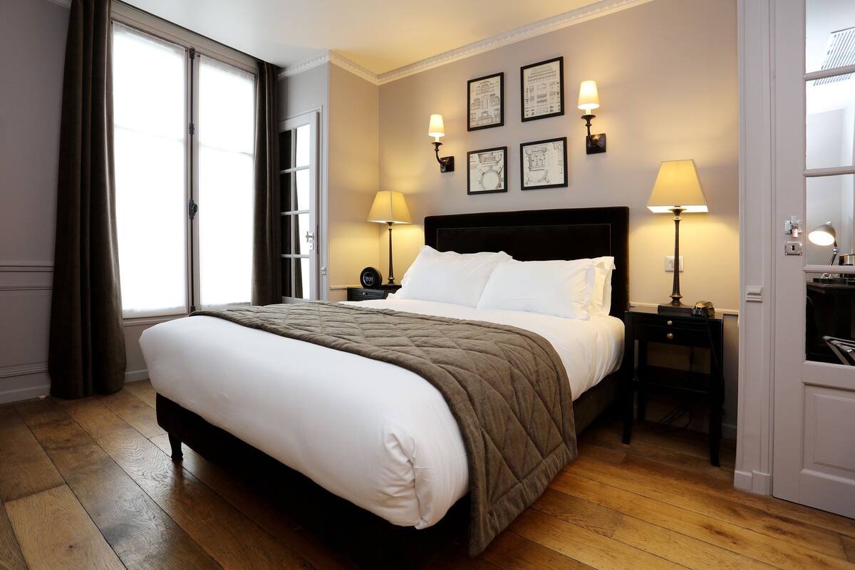 Una cama bien tendida con colcha, dos mesitas de noche con lámparas y un ventanal con cortinas.