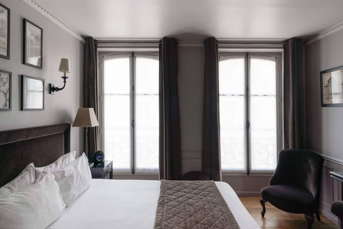 Habitación de hotel con una cama grande, una silla, una mesita de noche con lámpara y una ventana con cortinas.